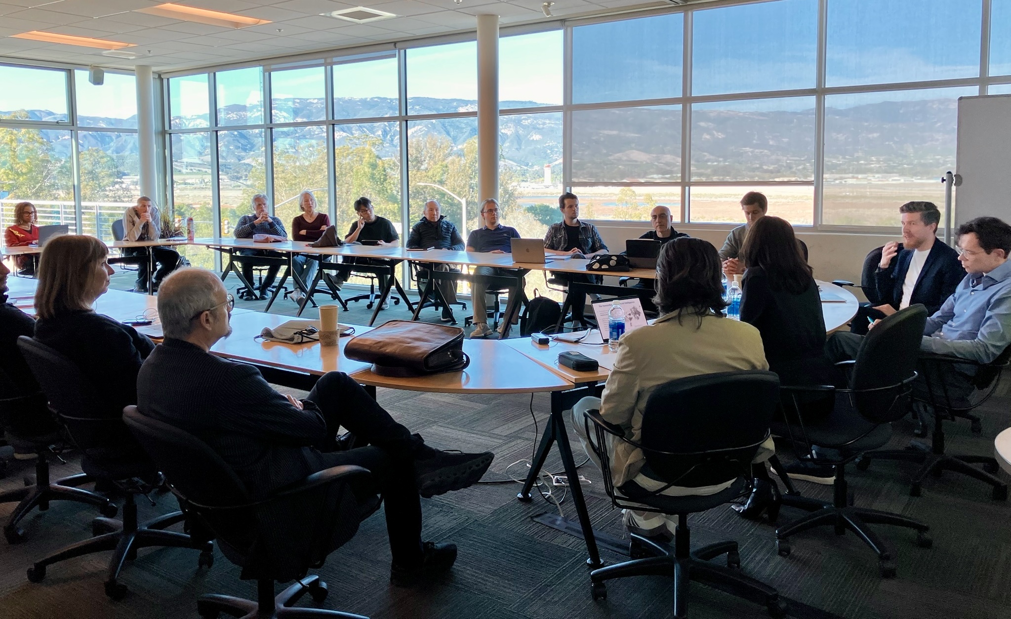 roughly twenty people sitting at a round table in a room with large windows through which a mountain range can be seen